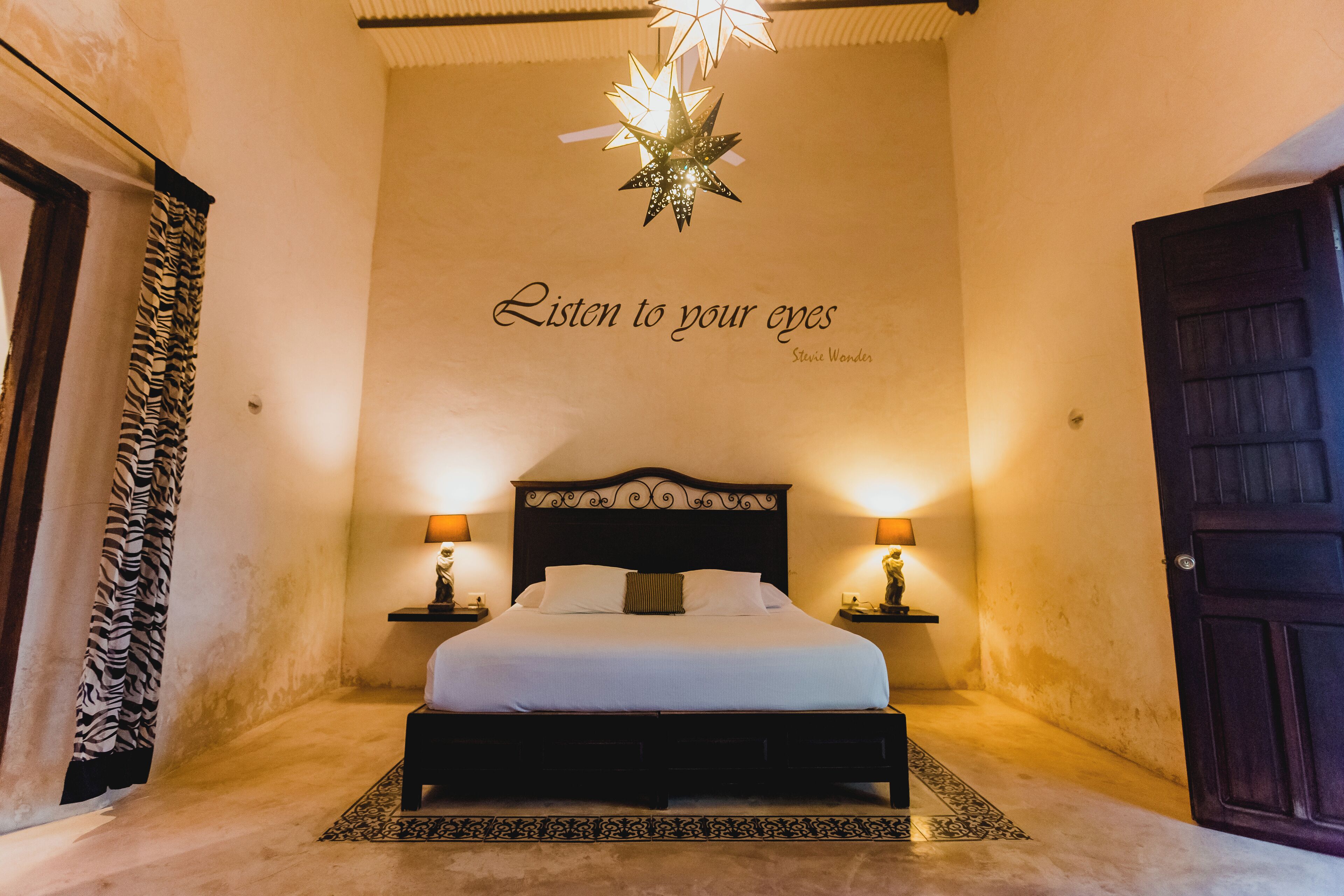 Une chambre à coucher avec un lit, deux tables de chevet avec des lampes, un rideau à motifs zébrés, un luminaire en forme d’étoile et un mur sur lequel est inscrit le texte « Écoutez ce que vos yeux vous disent », de Stevie Wonder.
