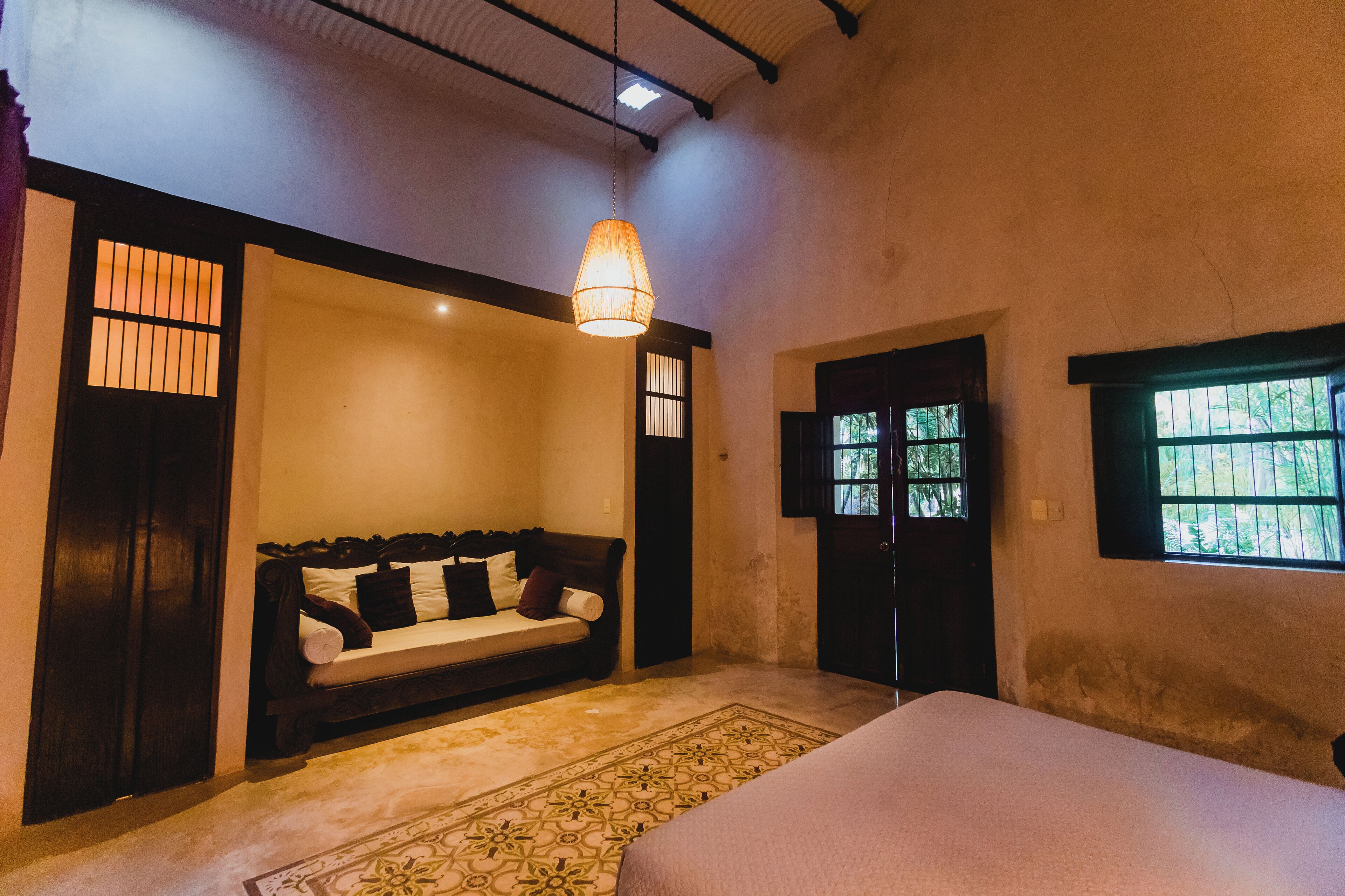A room with a bed, a sofa, a patterned rug, a hanging light fixture, and a window with bars.
