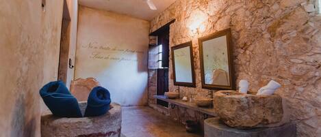 Un baño con paredes de piedra, un ventilador de techo, dos lavabos de piedra y un espejo.