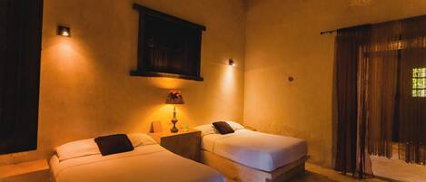 A room with two beds, a small table with a lamp, a wall-mounted cabinet, and a large window with curtains.