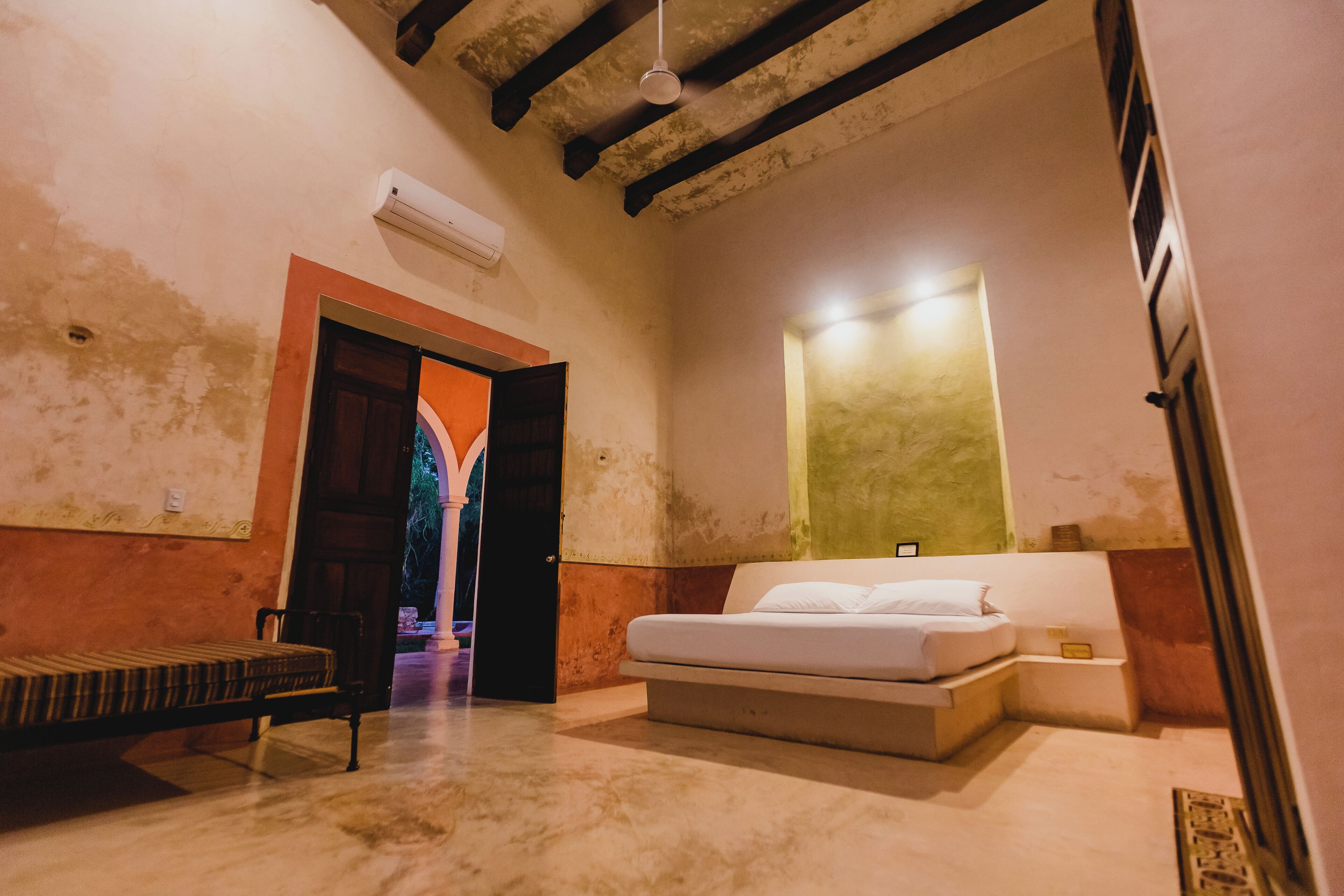 A bedroom with a bed, a bench, an open door leading to another room, and a wall-mounted light fixture.