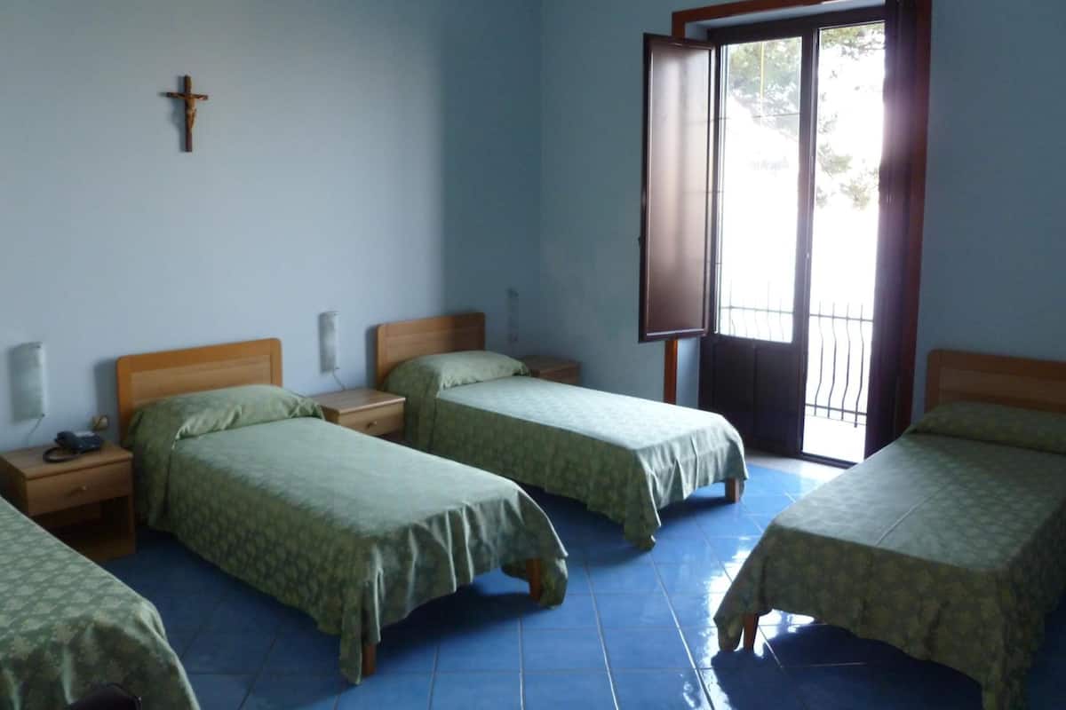 Een kamer met vier bedden, een kruis aan de muur, een raam met uitzicht en een deur.