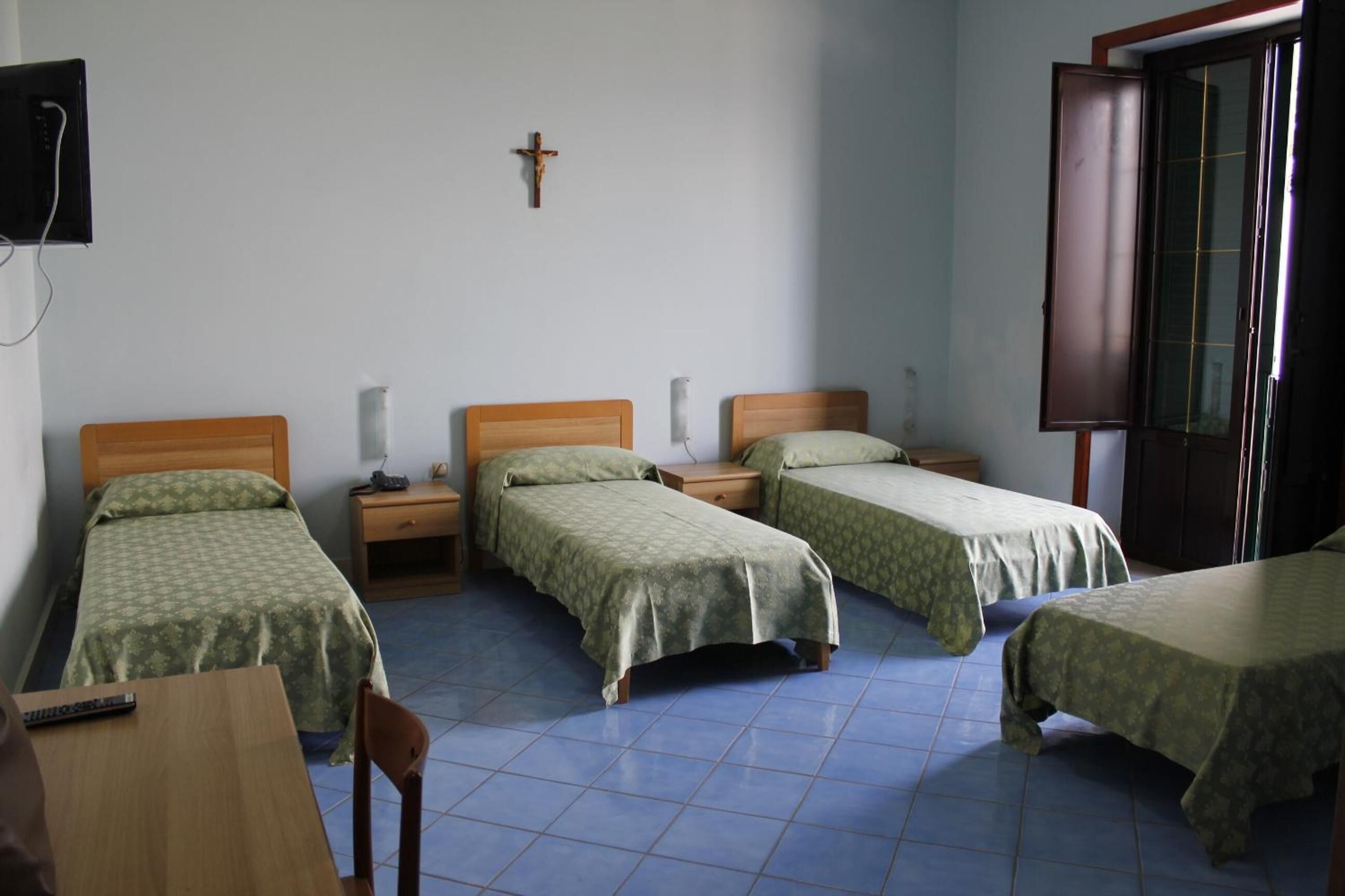 A room with four single beds, a wooden desk, a chair, a television on the wall, and a cross on the wall.