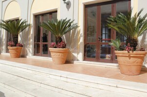 Un edificio con amplios ventanales y puertas, plantas en macetas y un área de entrada con pavimento de baldosas.