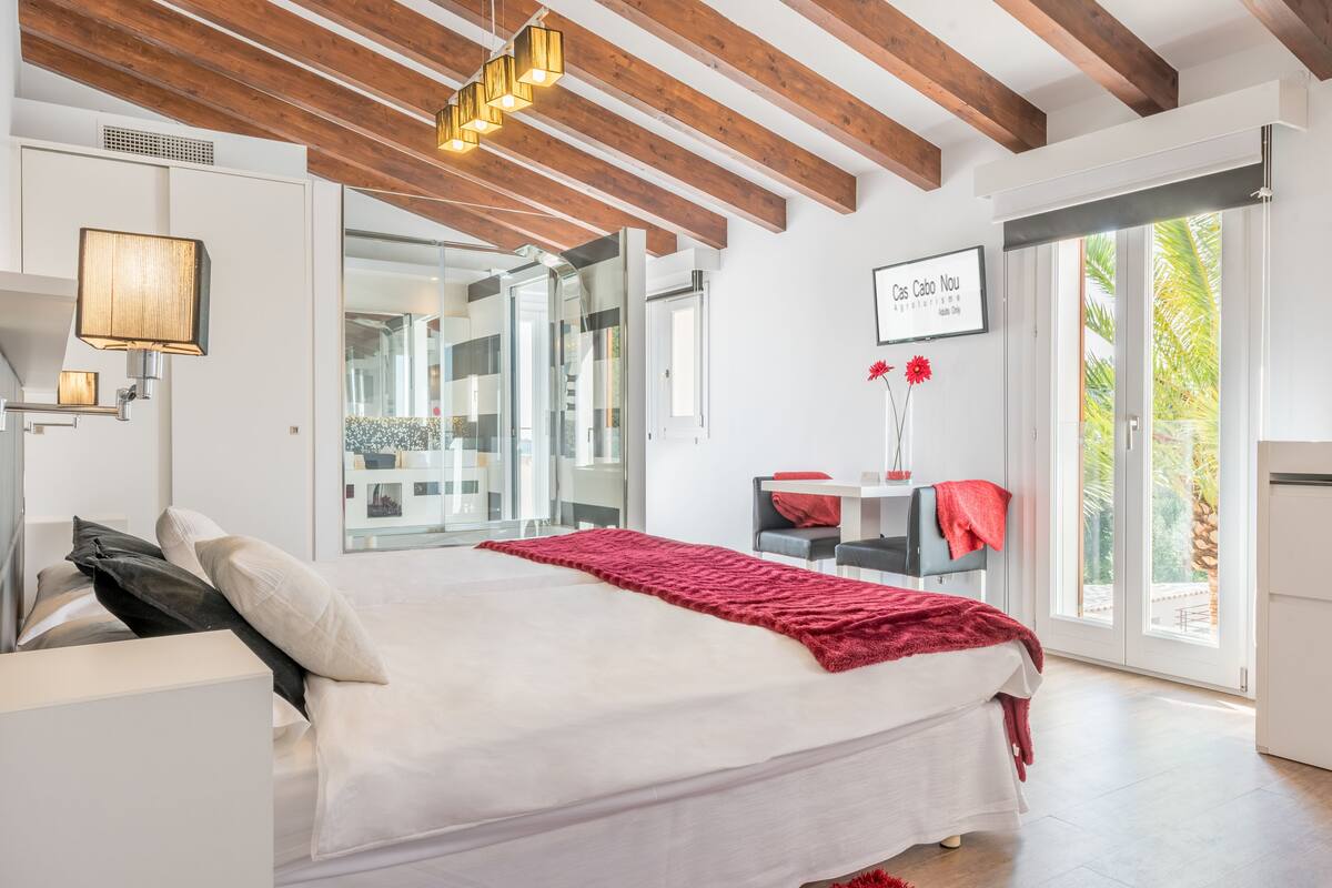 Une chambre moderne avec un grand lit, des poutres en bois, une porte vitrée donnant sur un balcon et une vue sur des palmiers.