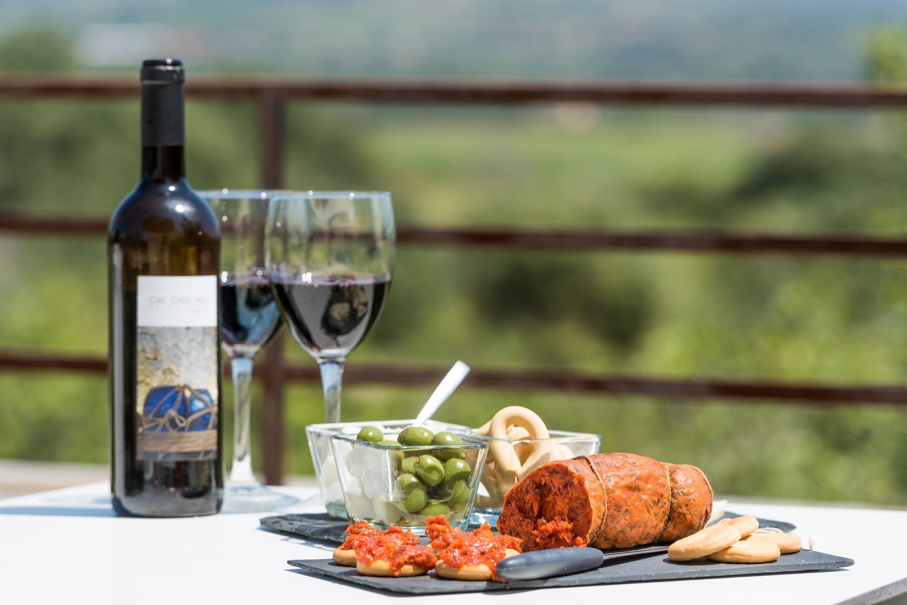 Une table avec une bouteille de vin, deux verres, des olives, une portion de fromage et des crackers, le tout installé sur une terrasse offrant une vue panoramique.