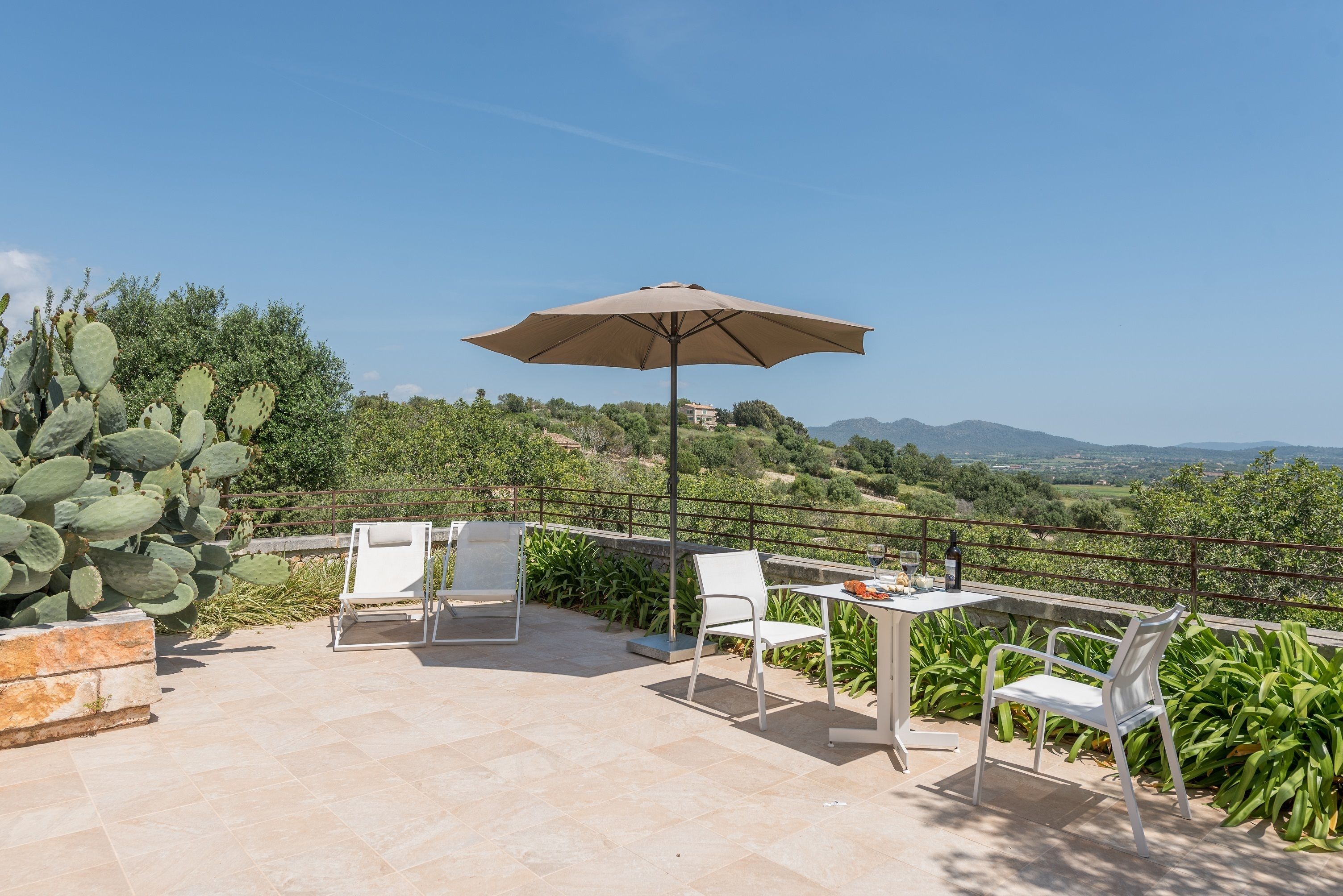 Un patio al aire libre con mesa y sillas, un paraguas color beige y vistas a un paisaje montañoso.