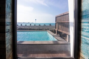 A view of a swimming pool with a wooden deck and a stone wall, overlooking a body of water and a distant cityscape. - Aqua BEAutique (Jeju City)