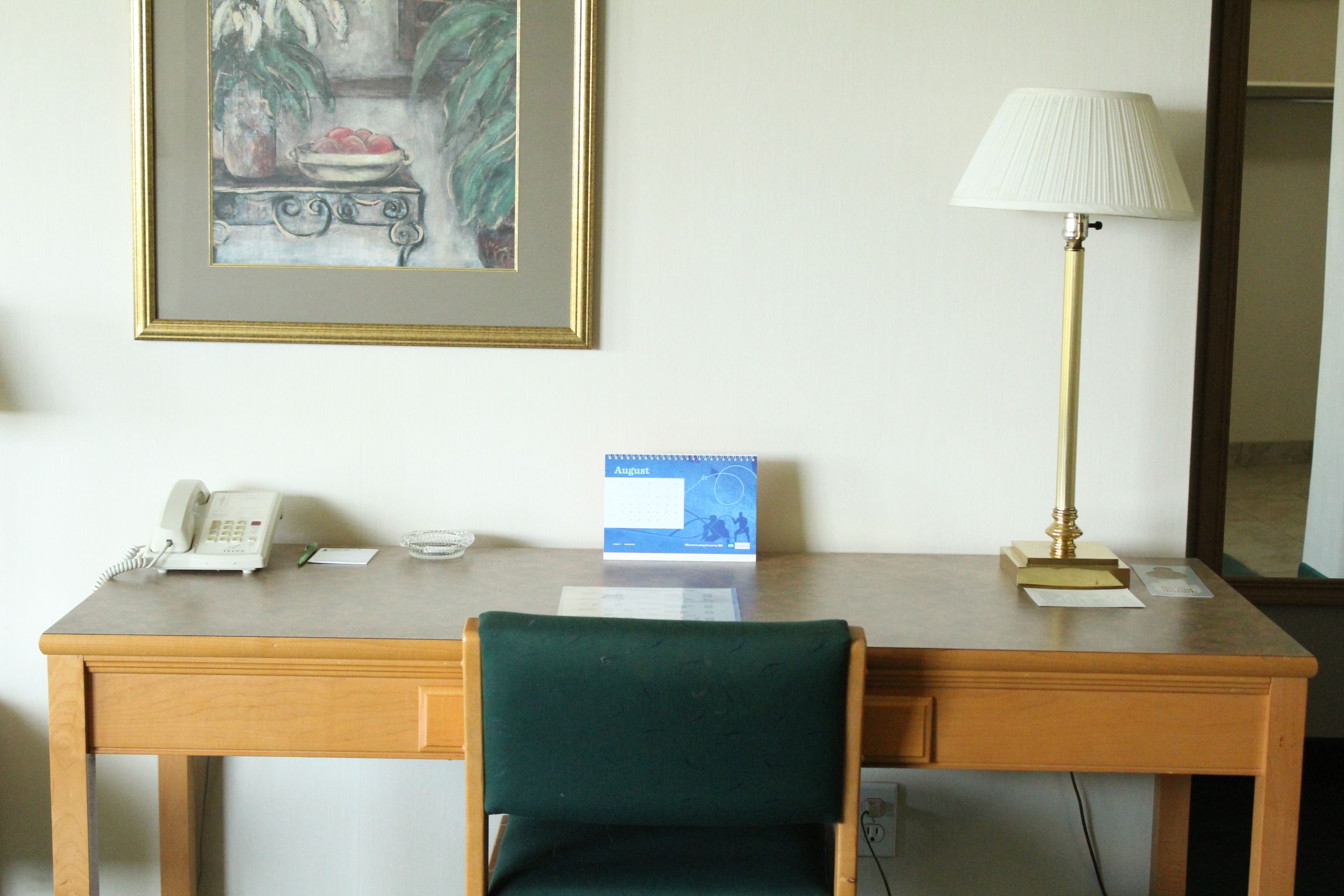 Habitación de hotel con escritorio de madera, una silla verde, una lámpara, un cuadro enmarco, un calendario y un teléfono.