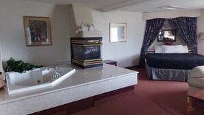 A hotel room with a large bathtub, a bed with dark bedding, a bedside table, a mirror, and a window with curtains. - Tamarack Motor Inn (Rocky Mountain House)