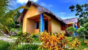Un bungalot tropical amb teulada de palla, envoltat de vegetació exuberant i palmeres.