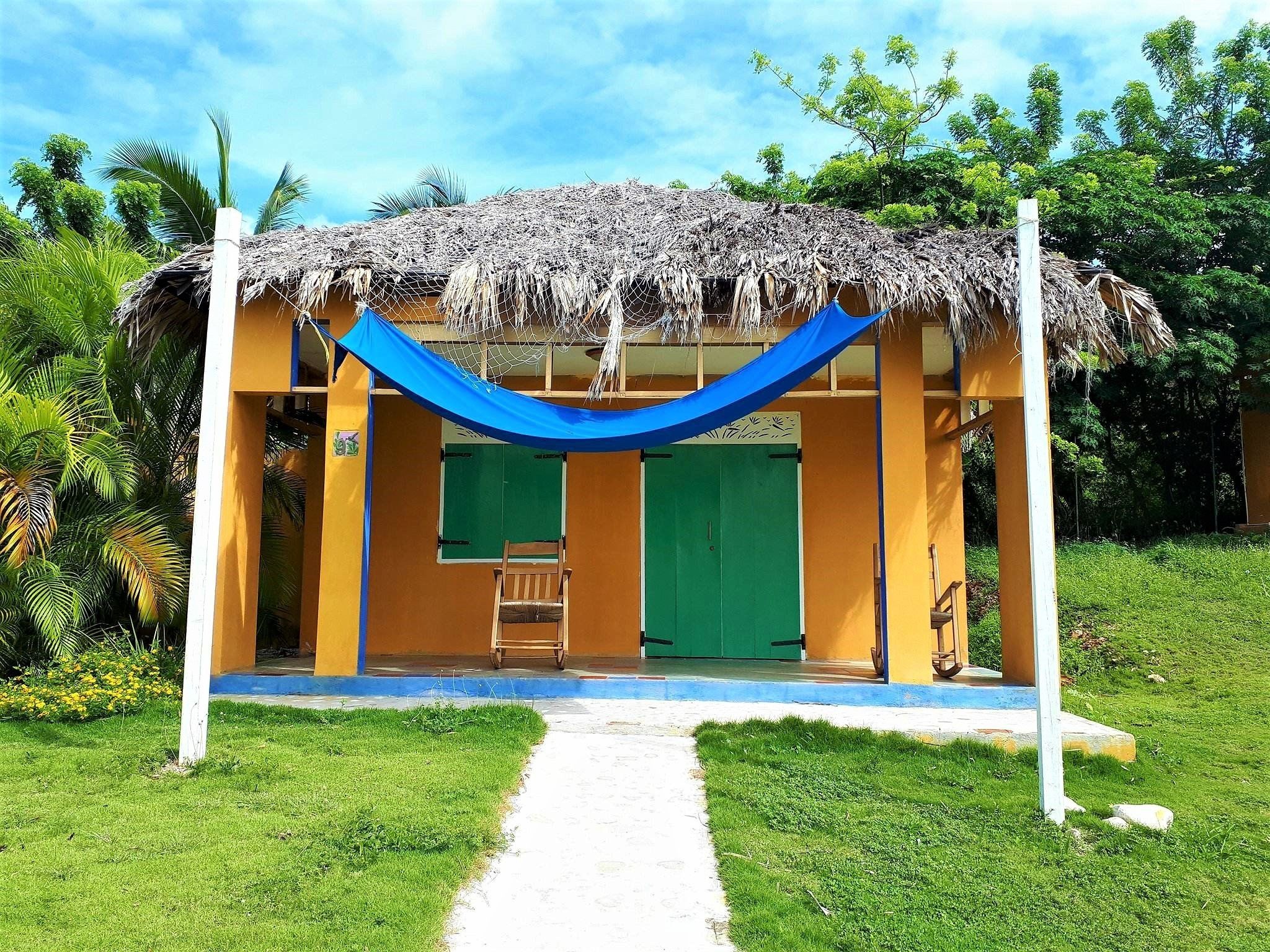 A thatched-roof structure with a blue canopy, a green door, and a bench.
