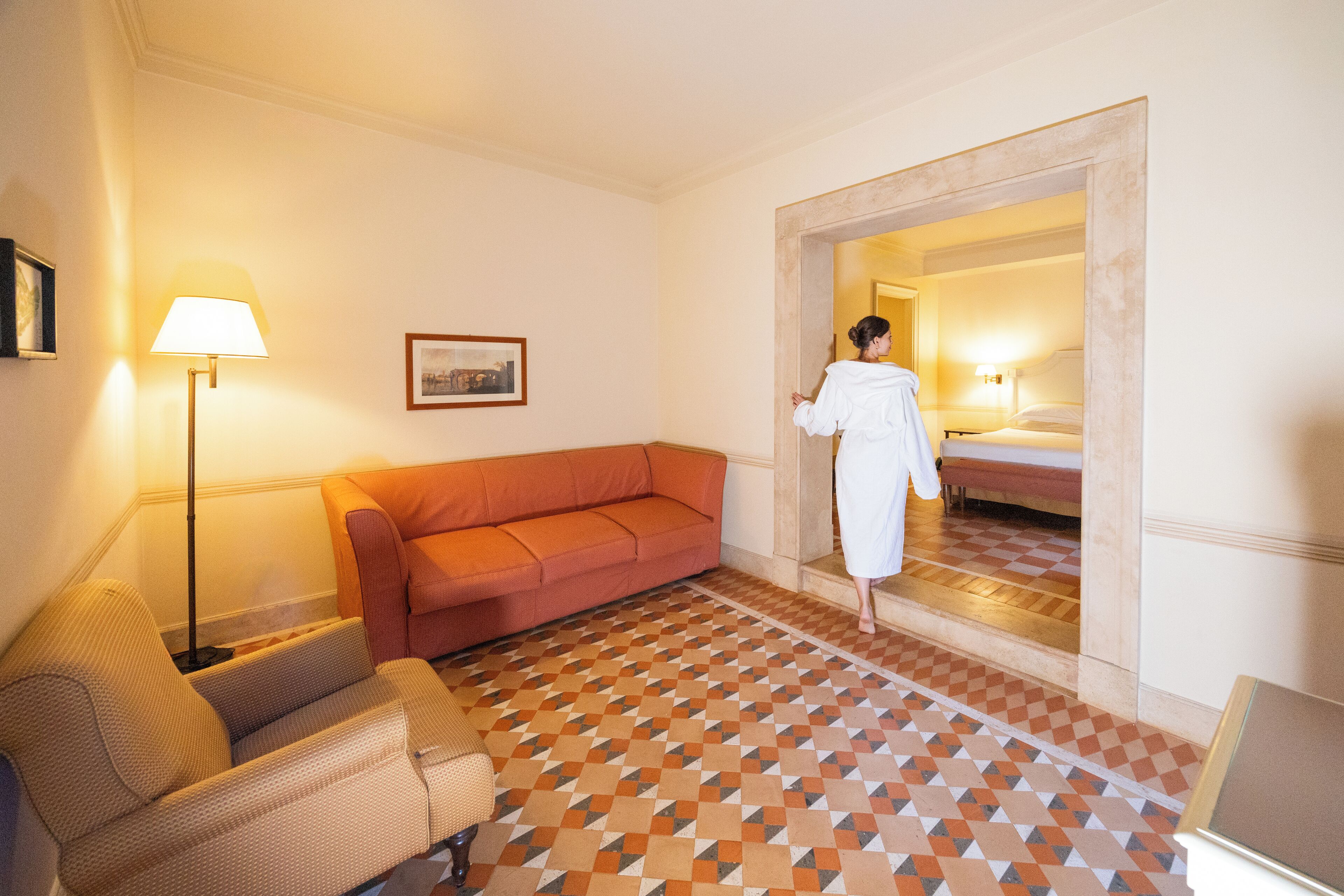 A hotel room with a checkered floor, a sofa, a lamp, and a bed with a nightstand.