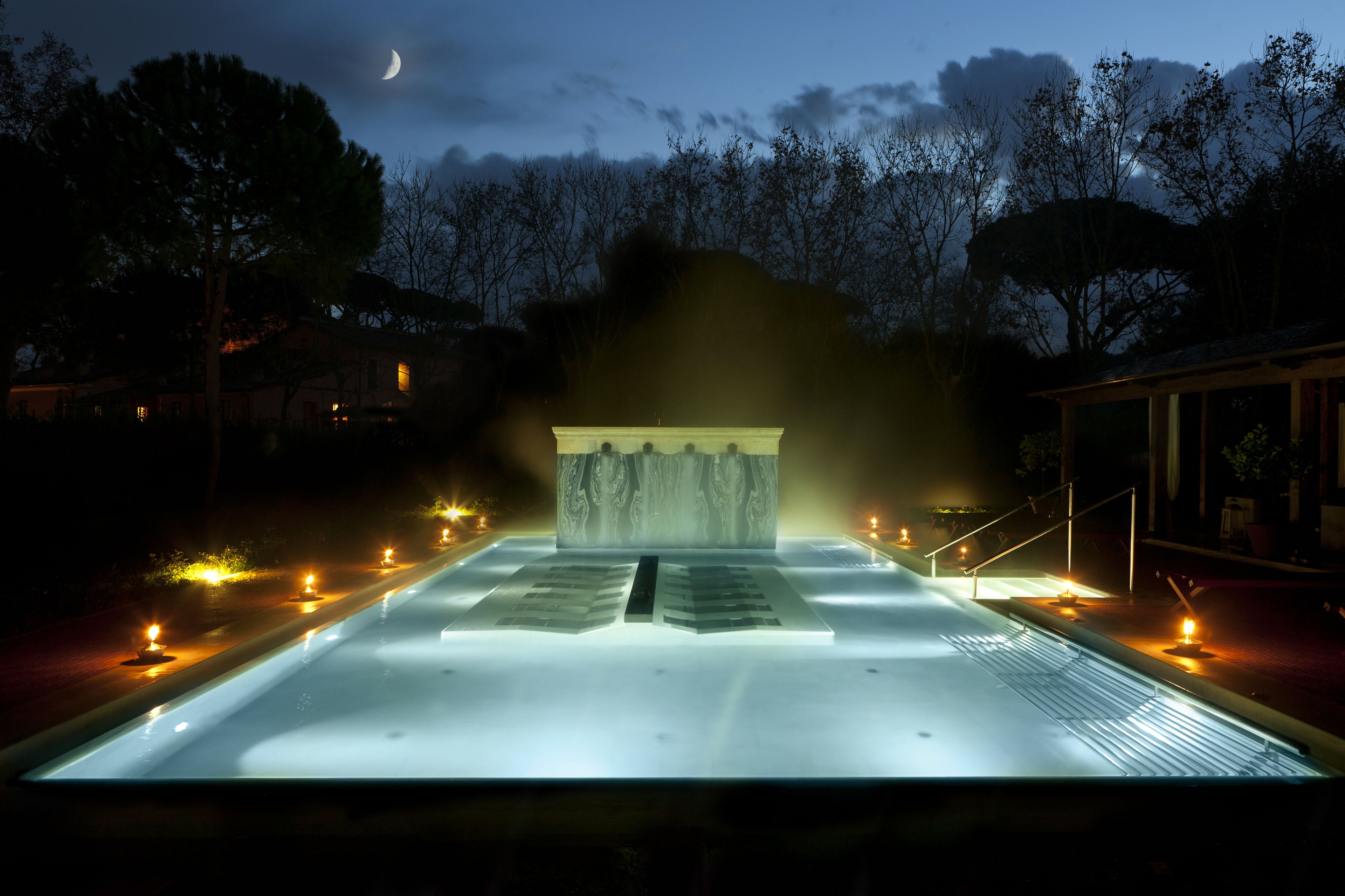 A serene outdoor pool area at night with a moonlit sky, a stone structure in the center, and a pathway lined with lights.