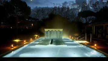 Un espace piscine extérieur paisible, éclairé la nuit par la lumière de la lune, avec une structure en pierre au centre et un chemin bordé de lampes.