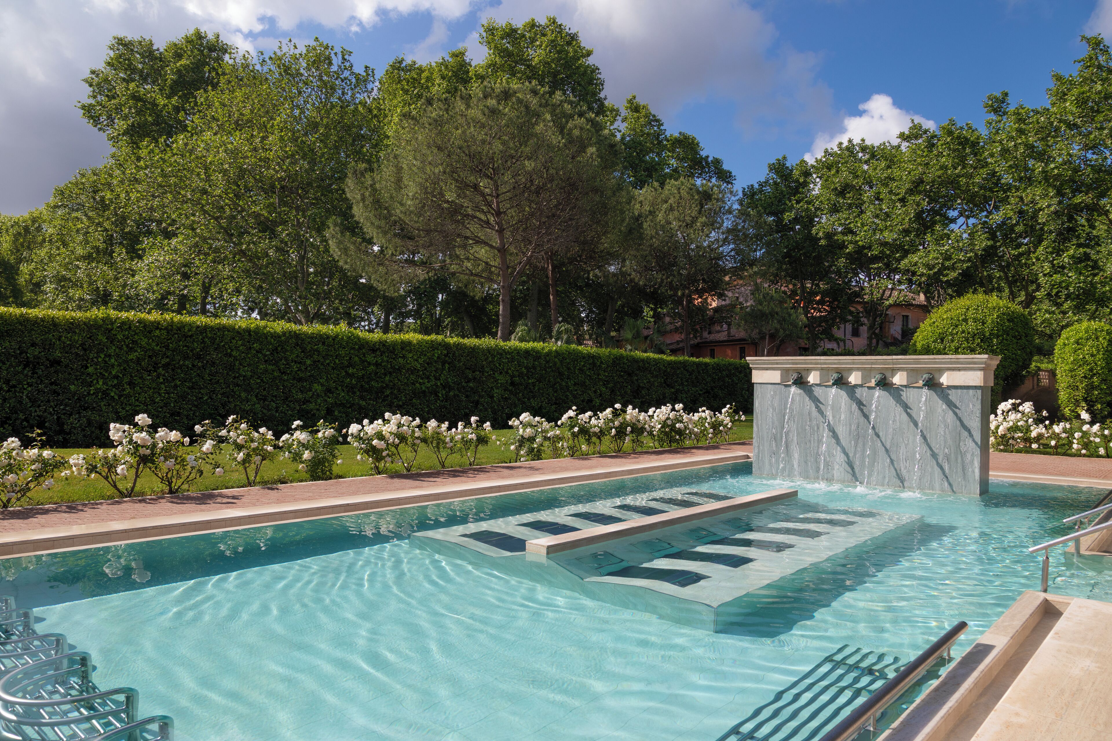 A swimming pool with clear water, surrounded by greenery and a hedge.