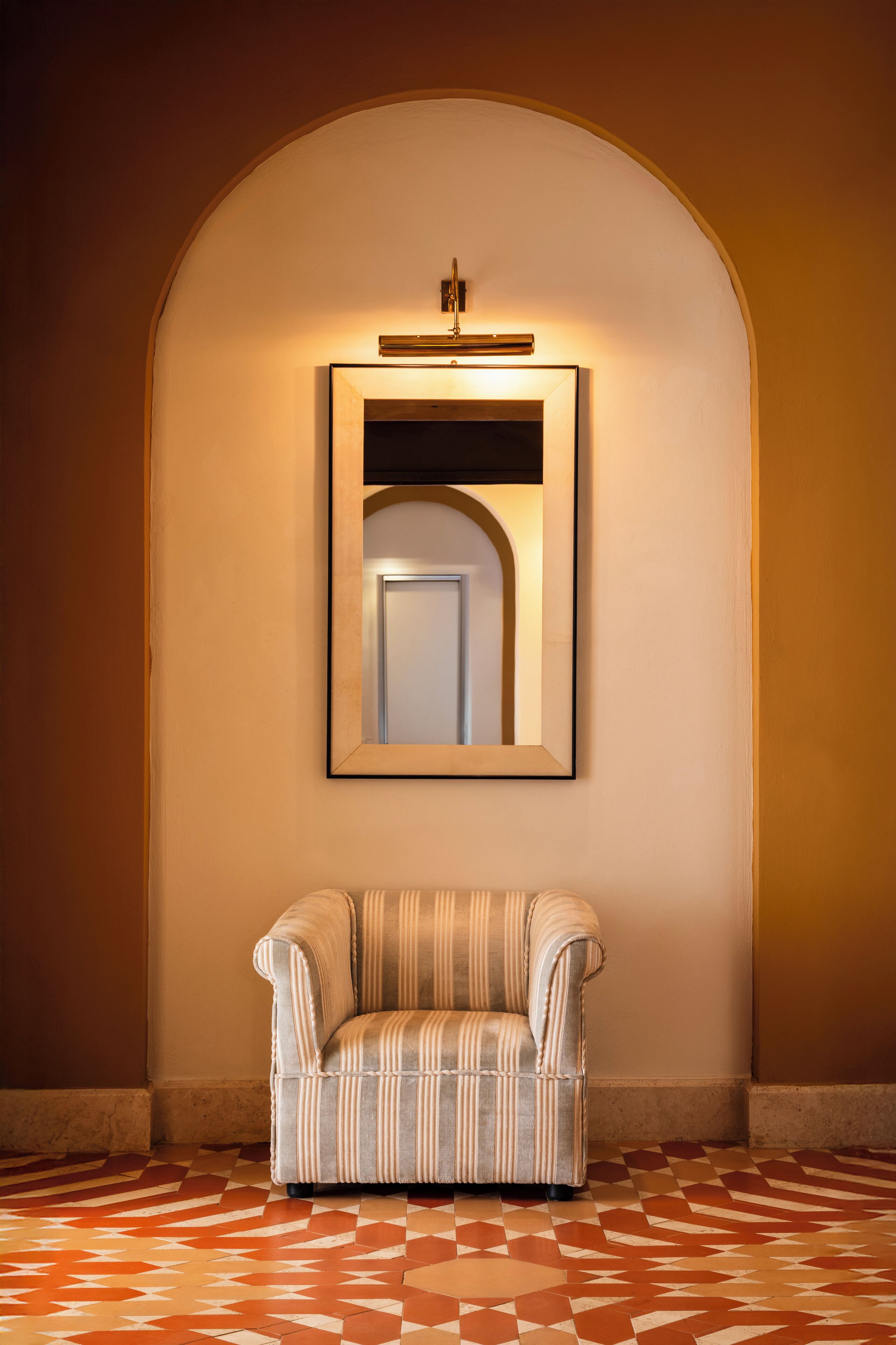 A chair in a hallway with a patterned floor and a framed mirror on the wall.