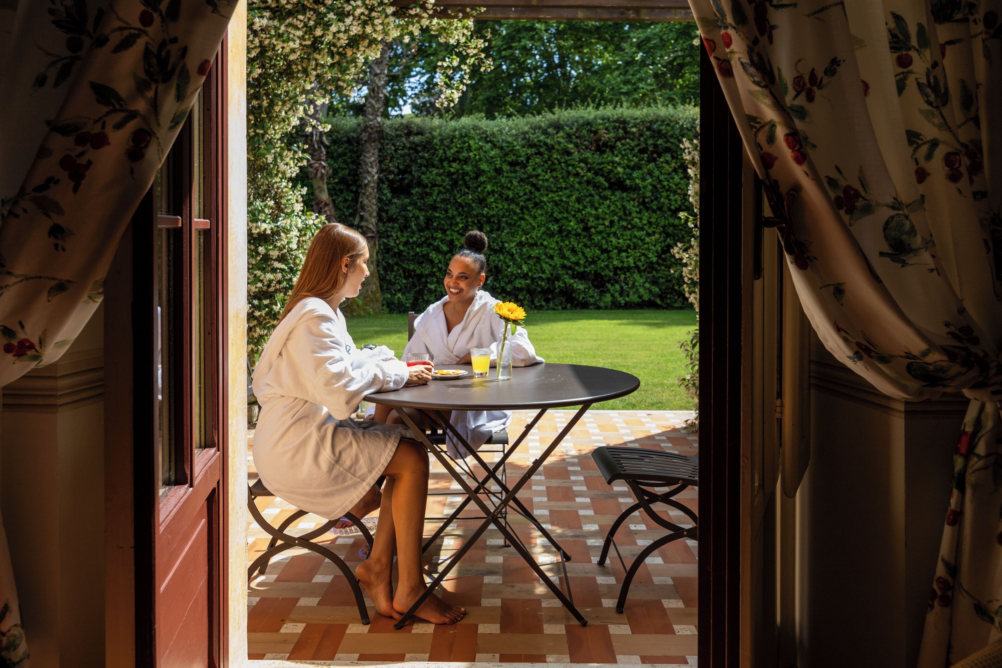 Une terrasse extérieure avec une table ronde et des chaises, une vue sur un jardin et une haie en arrière-plan.