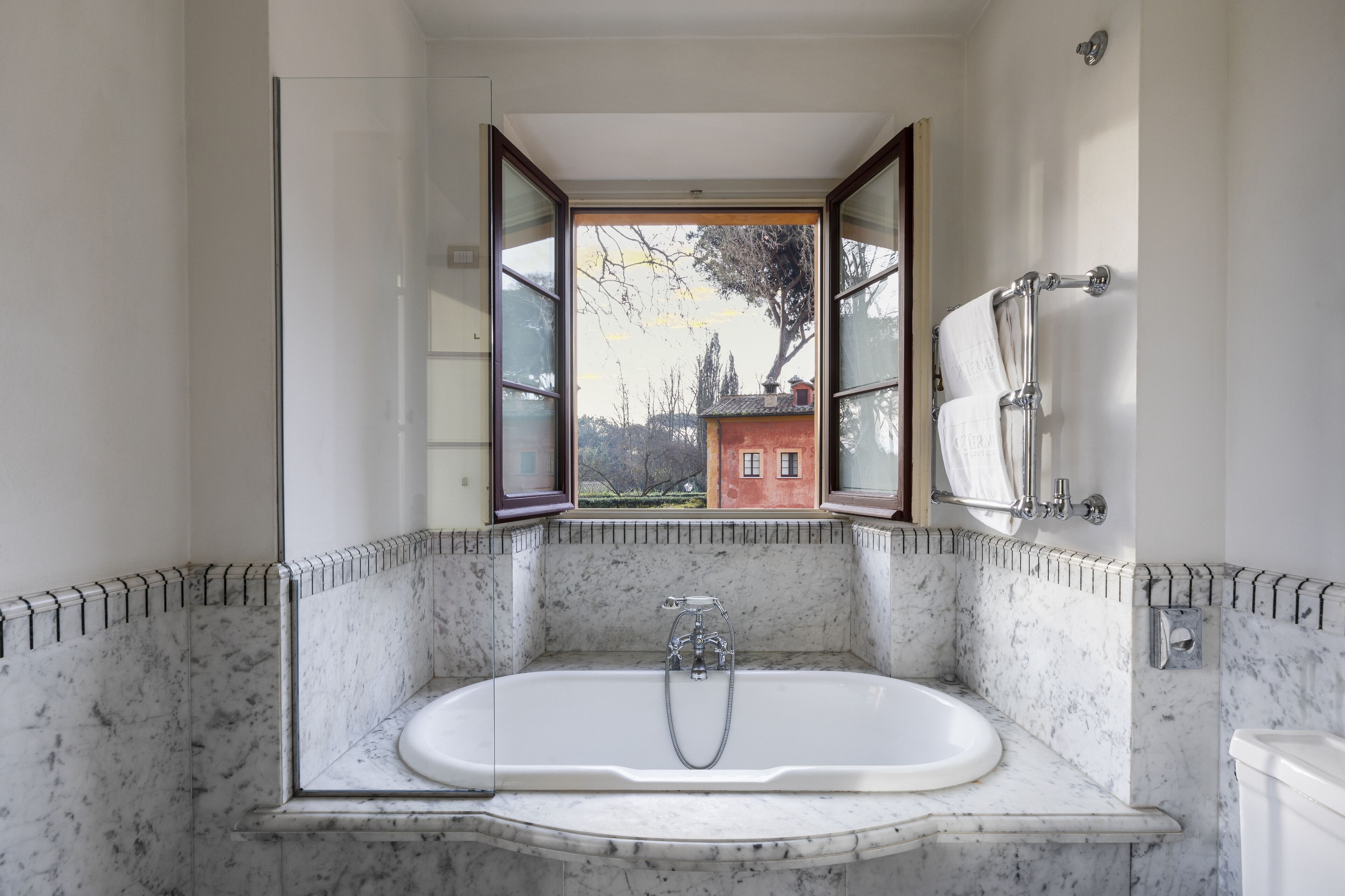 A bathroom with a large bathtub, a window with open shutters, and a view of a red building outside.