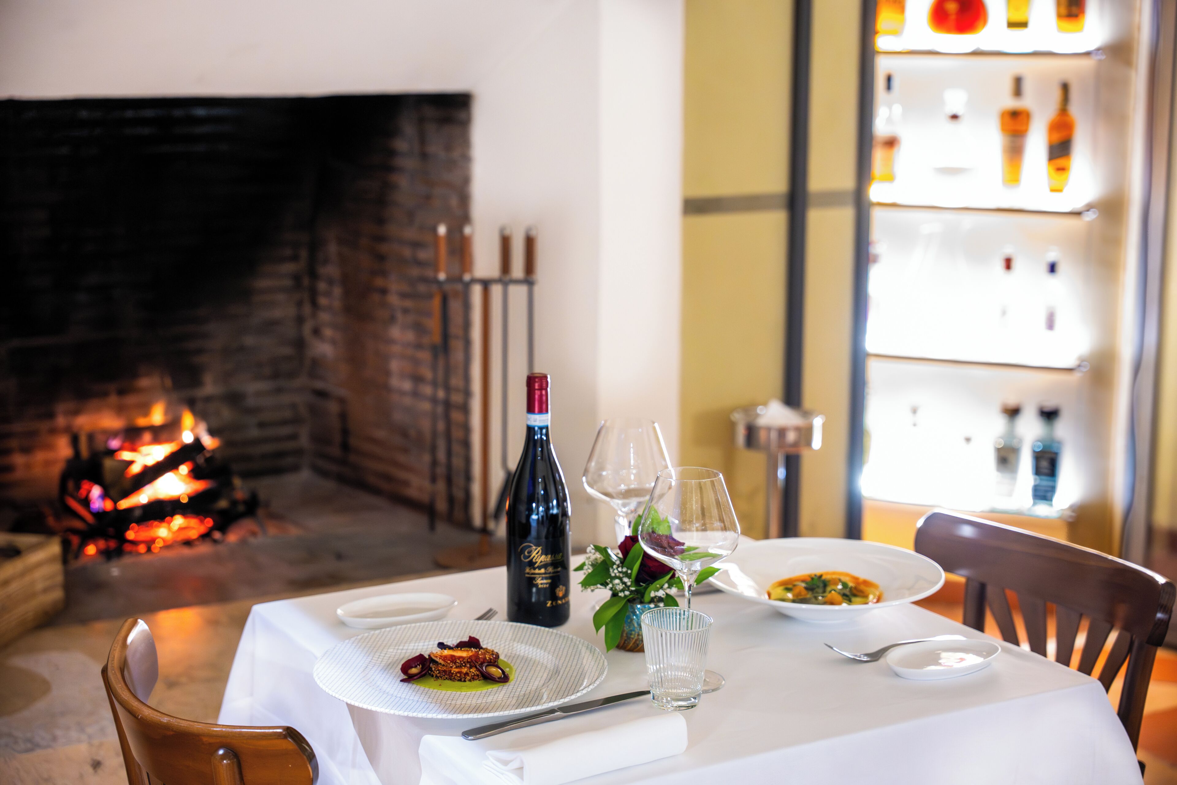 Une table à manger dressée avec des assiettes, une bouteille de vin, des verres et un vase rempli de fleurs.