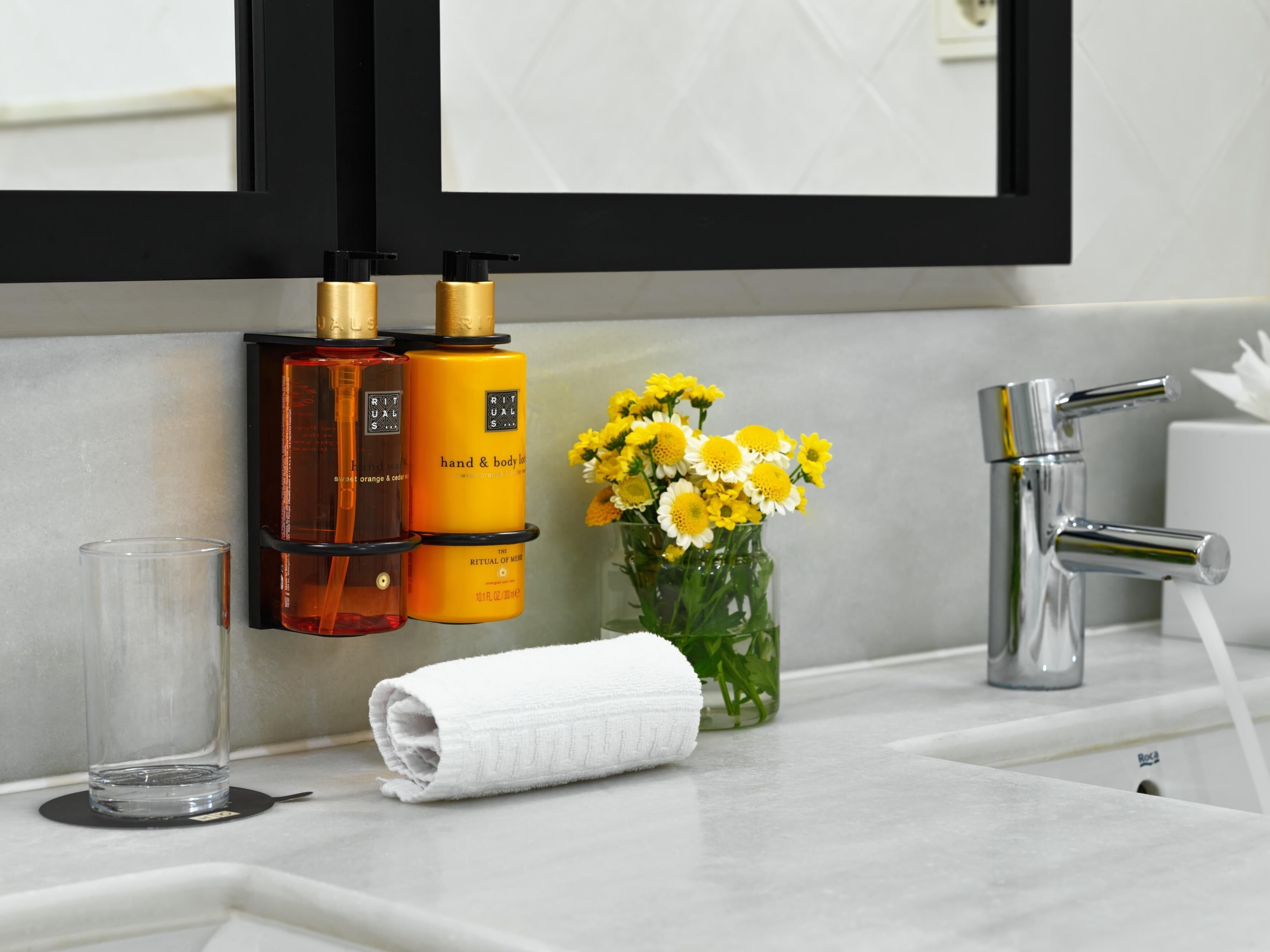 Un baño con espejo, dos botellas de crema para manos y cuerpo, un florero con flores amarillas, una toalla y un grifo.