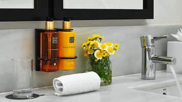Un baño con espejo, dos botellas de crema para manos y cuerpo, un florero con flores amarillas, una toalla y un grifo.