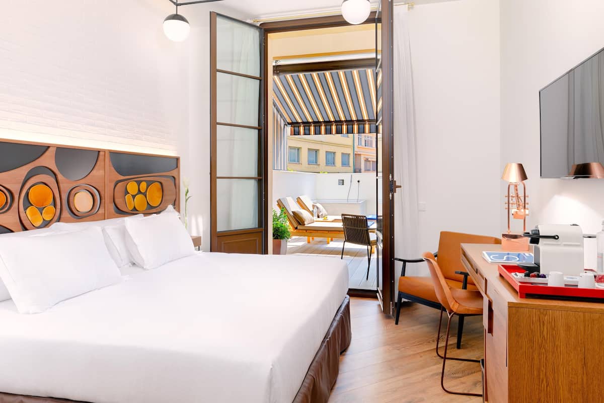 A hotel room with a large bed, a desk with a lamp, and a view of the city through an open door.