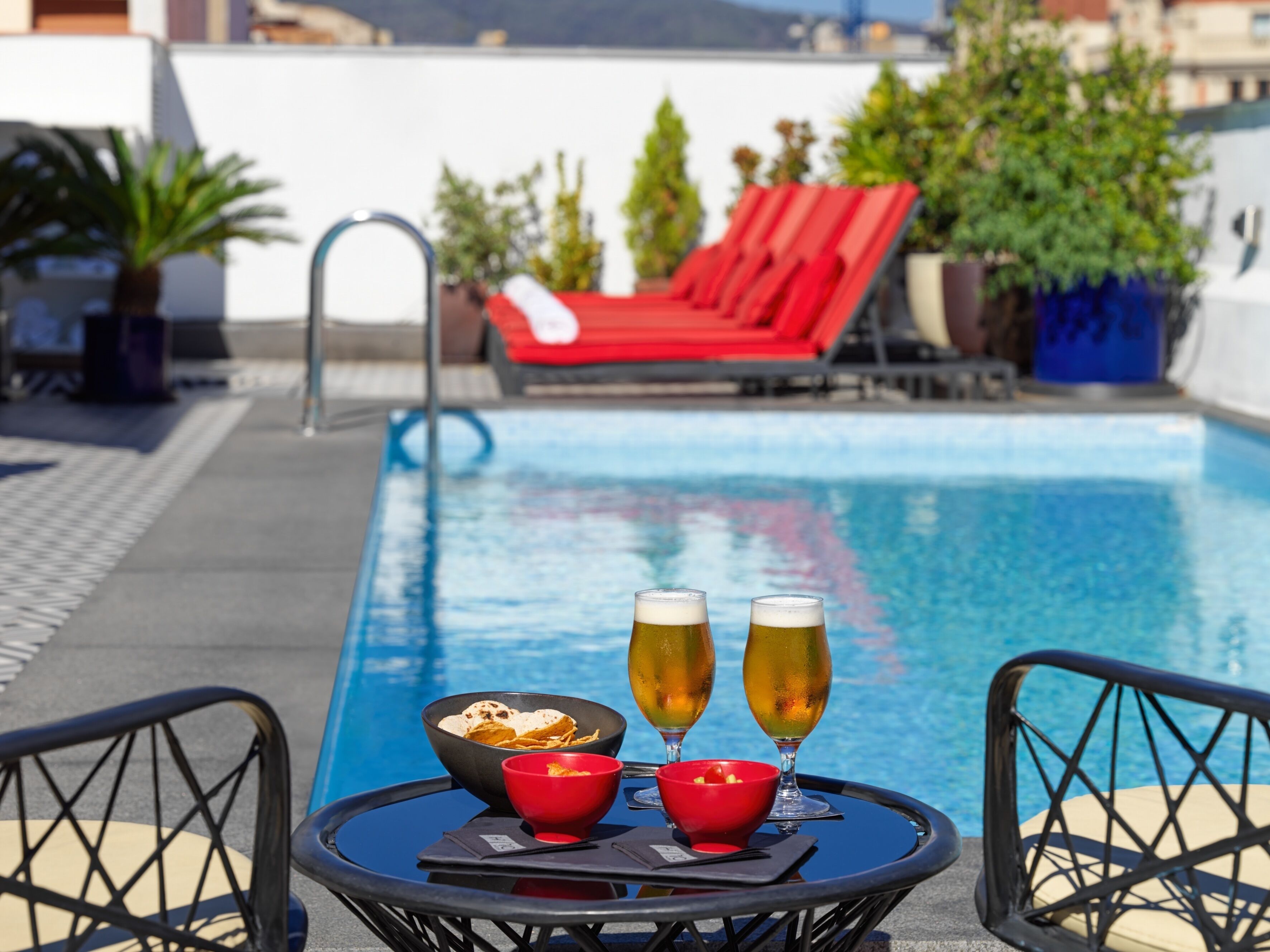 Mesa junto a la piscina con dos vasos de cerveza, un bol de patatas fritas y un bol de fruta.