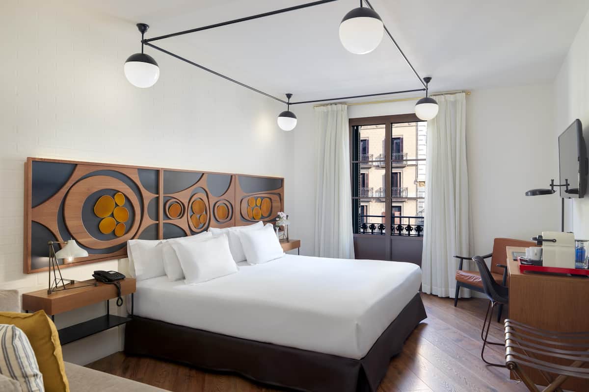 A modern hotel room with a large bed, a desk, a chair, and a view of the city through the window.