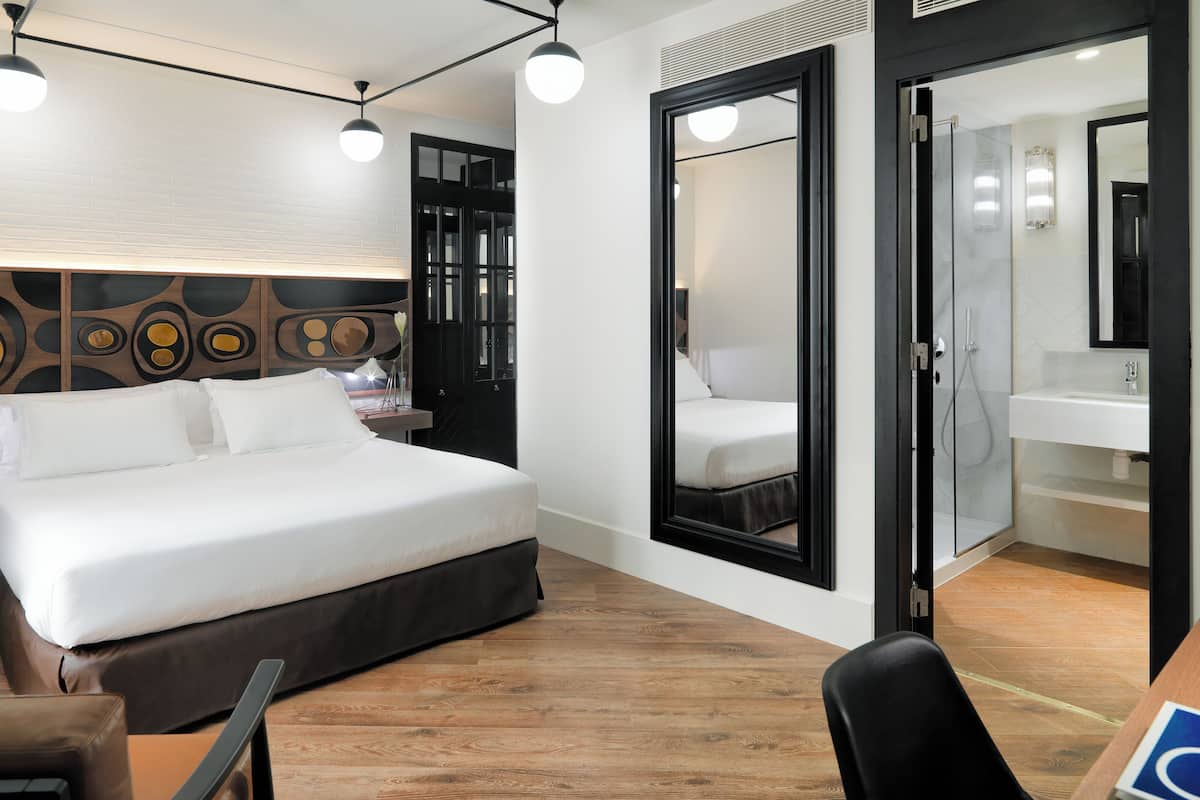 A modern hotel room with a large bed, a desk, a chair, a mirror, and a shower area.