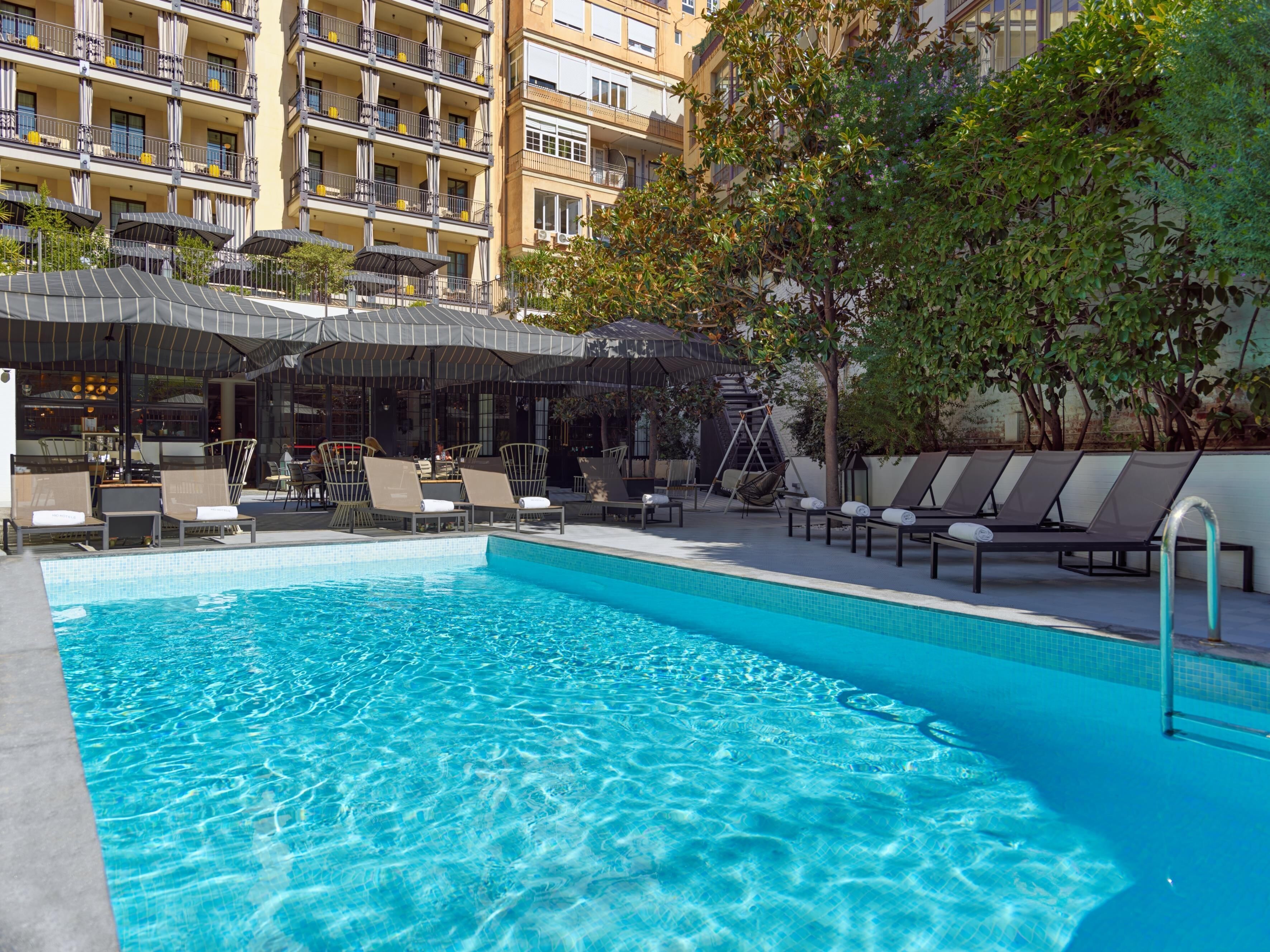 A swimming pool with lounge chairs and umbrellas, surrounded by a building with balconies and a tree.
