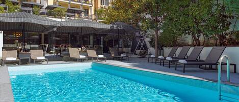 A swimming pool with lounge chairs and umbrellas, surrounded by a building with balconies and a tree.