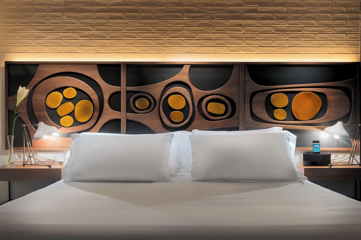 A hotel room with a large bed, two bedside tables, a modern wall art piece, and a nightstand with a lamp.
