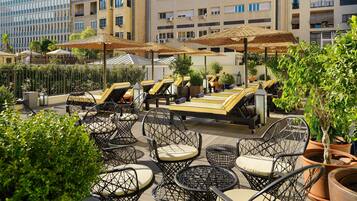 A rooftop terrace with outdoor seating, including chairs and tables, surrounded by potted plants and umbrellas.