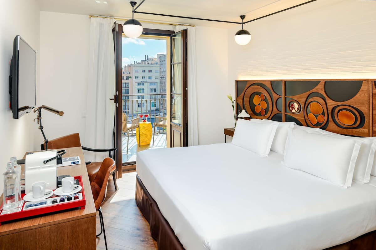 A hotel room with a large bed, a desk with a coffee machine, a chair, and a view of the city through the balcony.