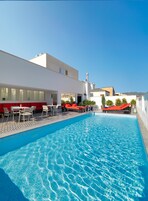 A swimming pool with a sun terrace, red lounge chairs, and a white building with a chimney.