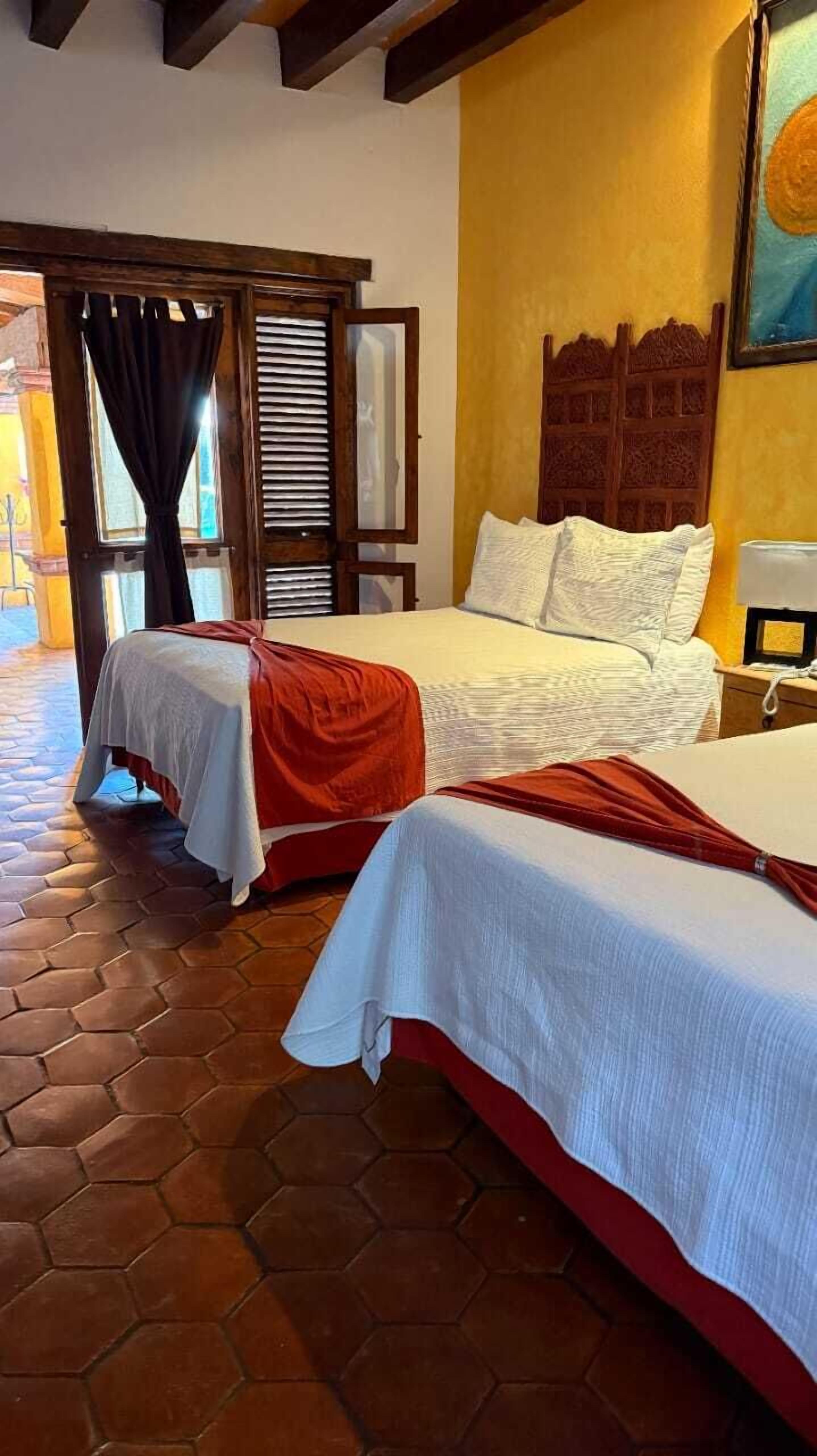 Habitación de hotel con dos camas, cabecera de madera, suelo de baldosas y una ventana con cortinas.