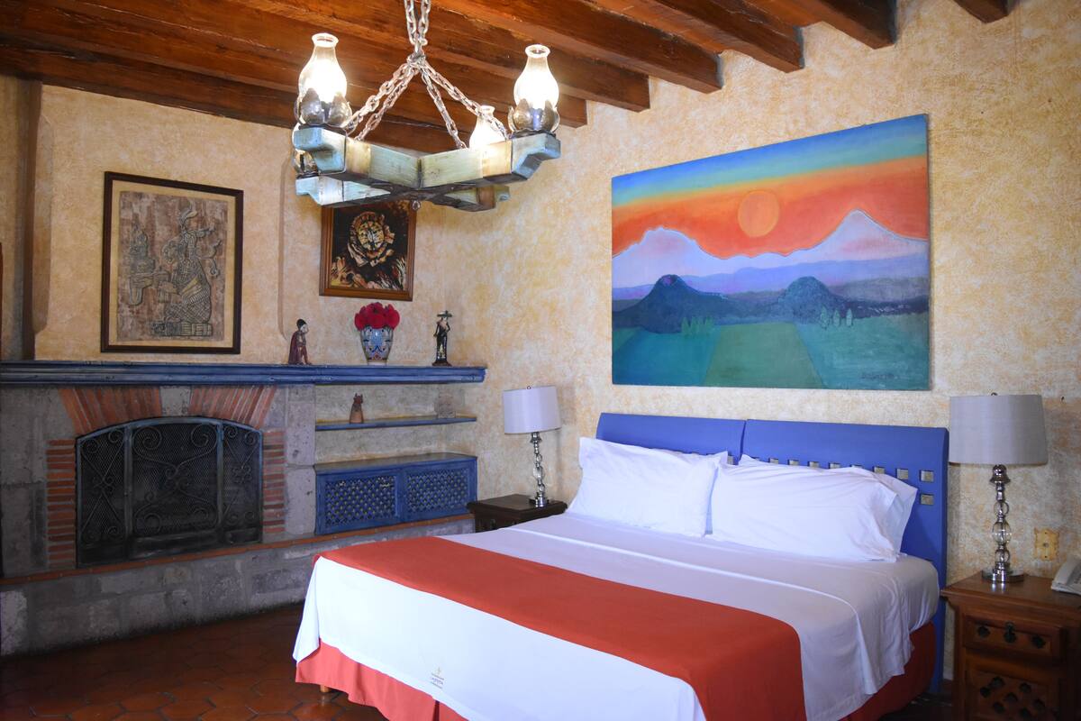 A bedroom with a bed, a fireplace, a painting, and a chandelier.