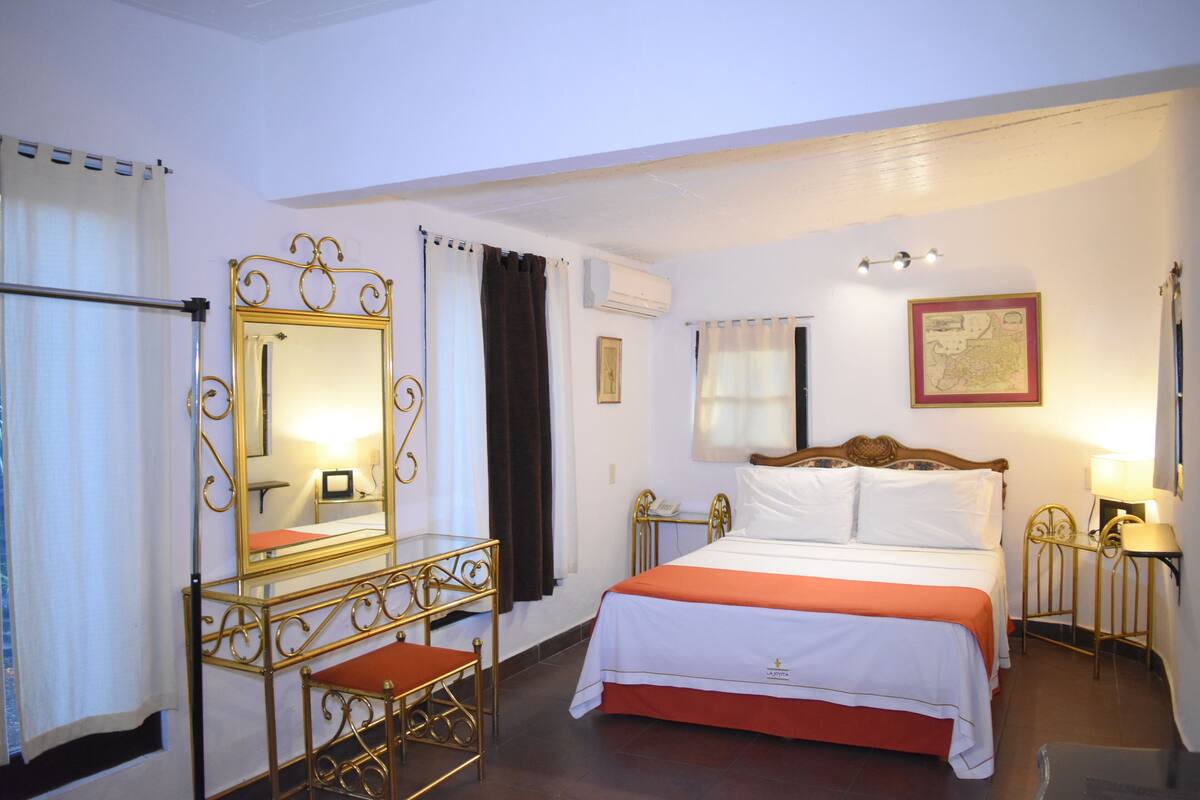 A hotel room with a bed, a vanity with a mirror, a small table, and a wall-mounted lamp.