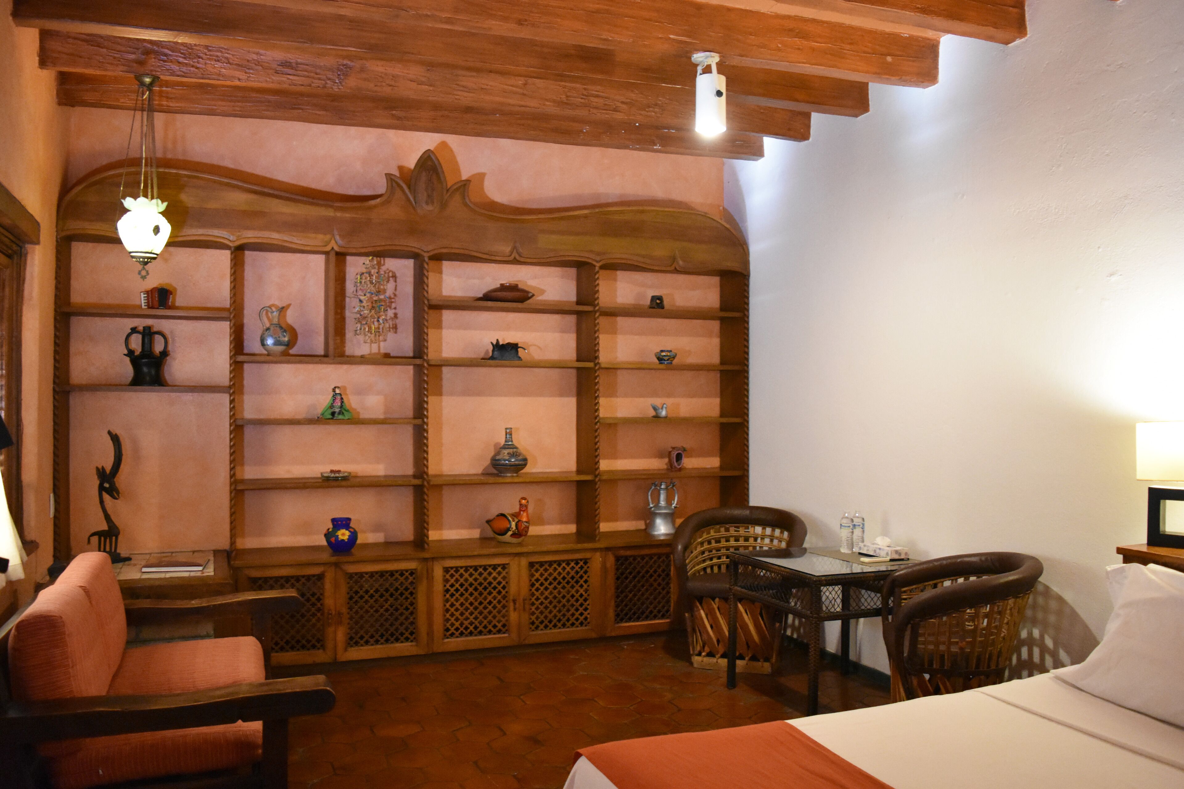 A cozy room with a bed, a wooden shelf with various decorative items, a wicker chair, and a small table with a lamp.