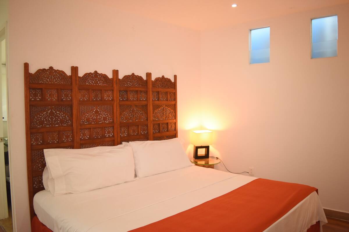 A neatly made bed with white linens and a red blanket, a wooden headboard with intricate carvings, a bedside lamp, and two small windows on the wall.