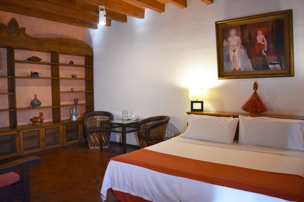 A hotel room with a bed, a desk with chairs, a shelf with decorative items, and a painting on the wall.