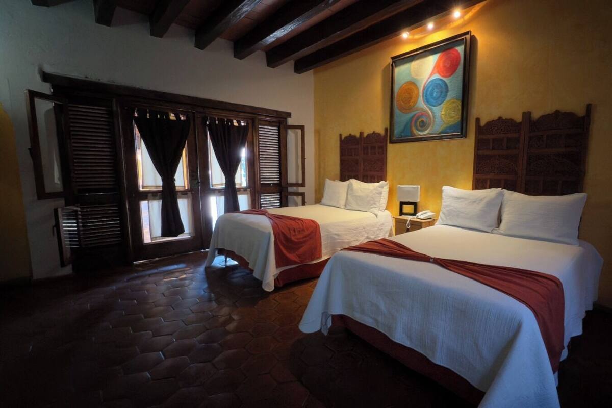 A hotel room with two beds, a large painting on the wall, and a window with shutters.