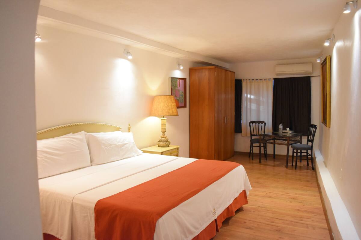 A hotel room with a bed, a table with chairs, an air conditioning unit, and a painting on the wall.