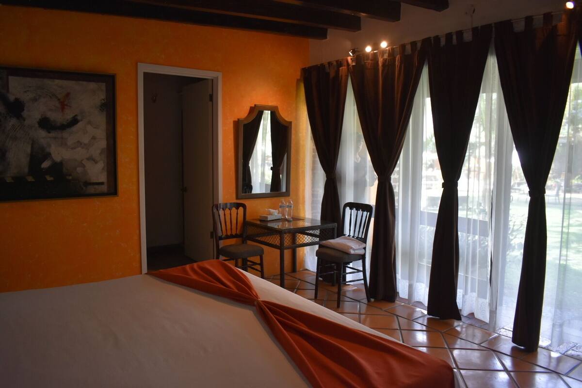 A hotel room with a bed, a desk, two chairs, a mirror, and a window with curtains.