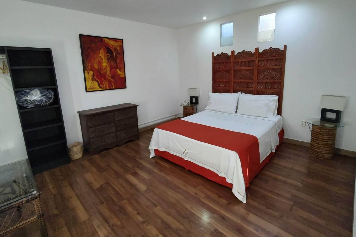 A bedroom with a wooden floor, a bed with white bedding, a wooden headboard, a red accent on the bed, a painting on the wall, a nightstand with a lamp, and a small table with a lamp.