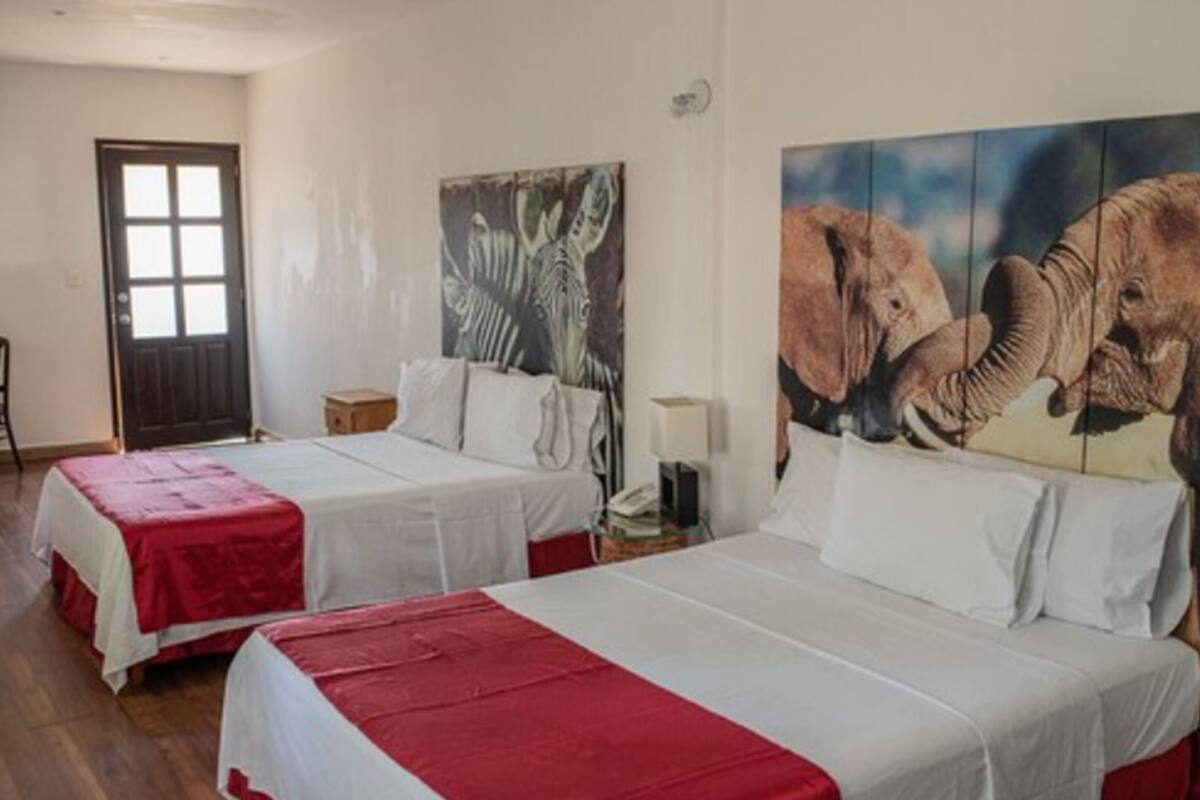 A hotel room with two beds, each with a red and white bedspread. There is a painting of elephants on the wall.