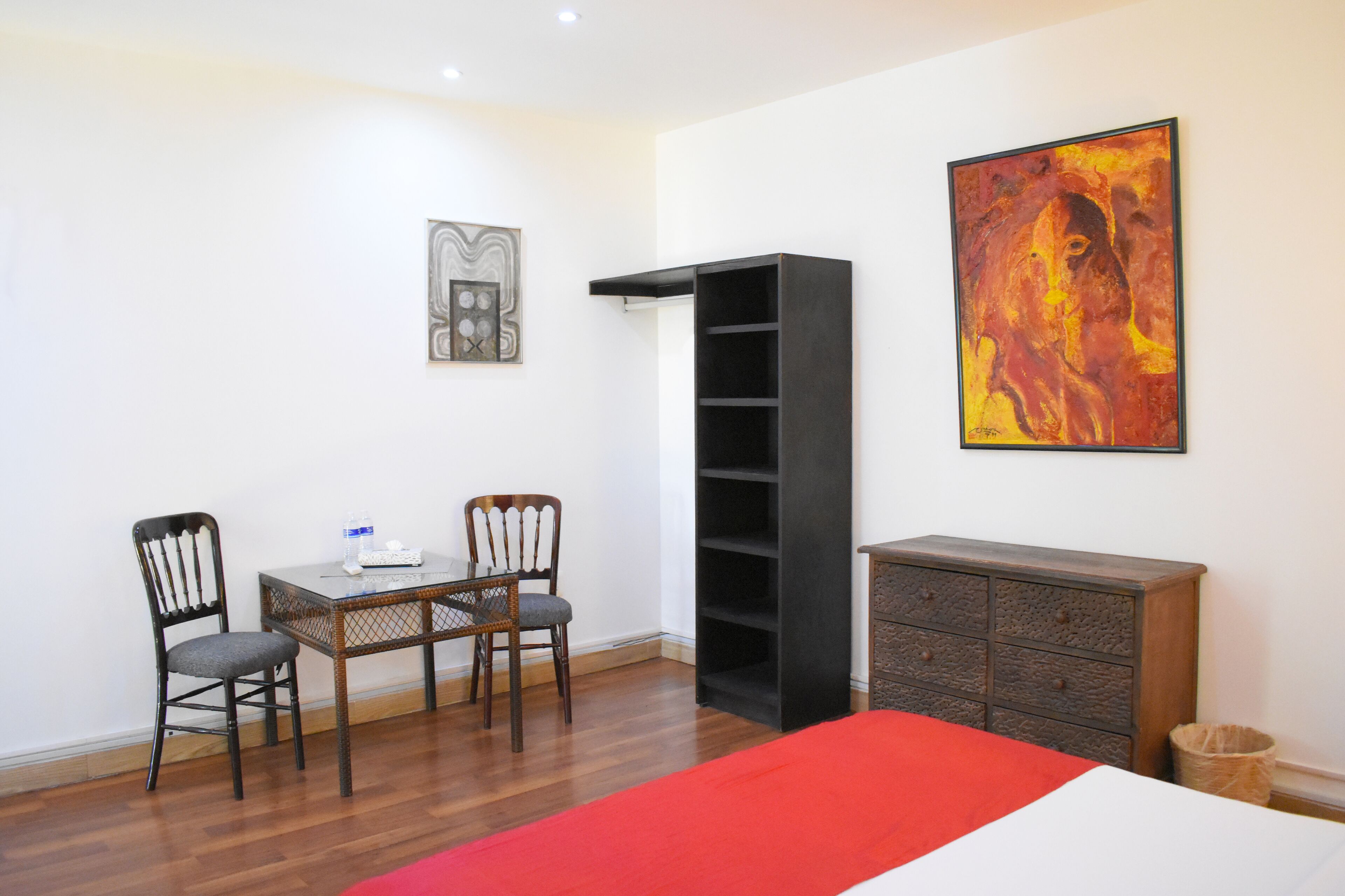 A bedroom with a bed, a desk with two chairs, a bookshelf, and two paintings on the wall.