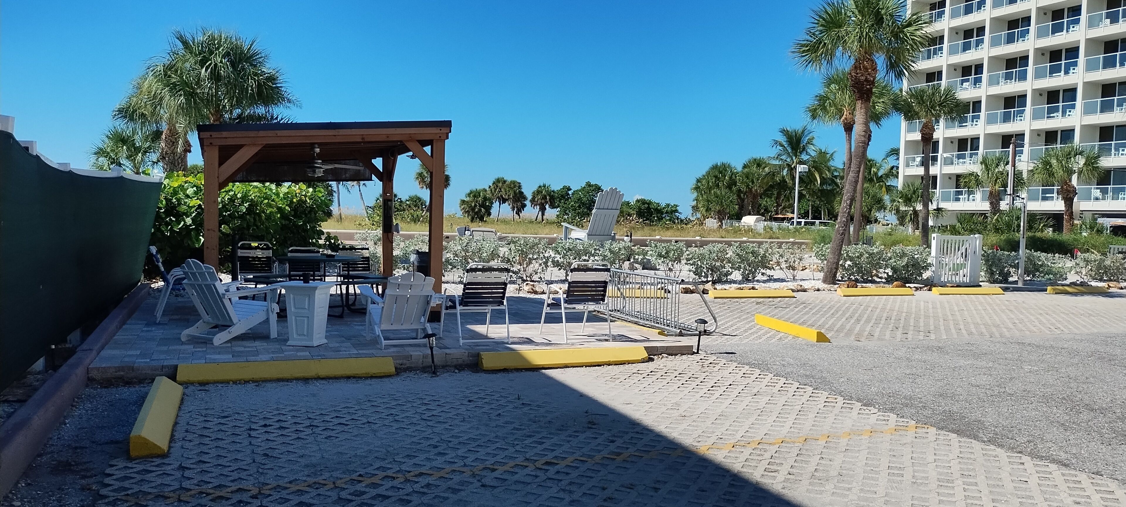 A covered outdoor seating area with chairs and tables, a paved parking lot, and a building with balconies in the background.