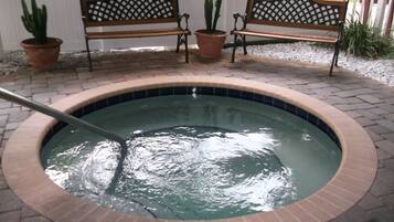 A hot tub with a tiled edge, surrounded by a brick patio. There are two wooden benches with lattice backs and potted plants nearby.
